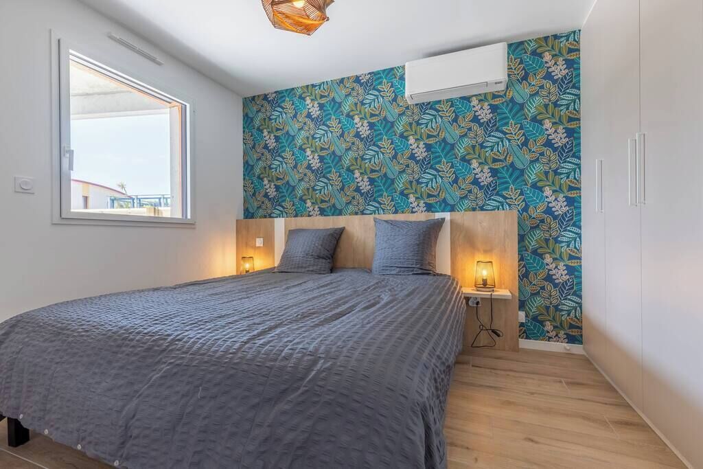 A bedroom with a bed, a window, a wall-mounted air conditioner, and a patterned wallpaper.