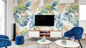 Un salon moderne doté d’un grand téléviseur à écran plat, de fauteuils en velours bleu, d’une table basse ronde et d’un mur orné d’un motif de feuilles tropicales.