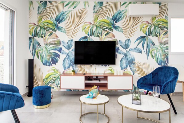 A modern living room with a large flat-screen TV, blue velvet armchairs, a round coffee table, and a tropical leaf-patterned wall.