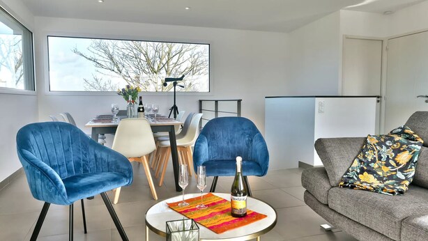 Un salon moderne avec un coin repas, comprenant des fauteuils en velours bleu, un canapé gris agrémenté d’un plaid à motifs floraux et une grande fenêtre donnant sur un paysage arboré.