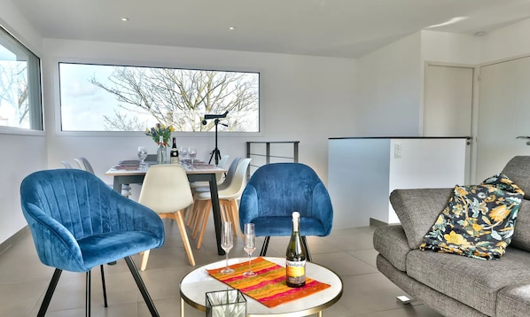 Un salon moderne avec un coin repas, comprenant des fauteuils en velours bleu, un canapé gris agrémenté d’un plaid à motifs floraux et une grande fenêtre donnant sur un paysage arboré.