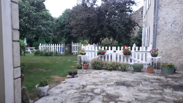 A stone-paved entrance with a white picket fence, potted plants, and a small table with a chair. - Appartement 4pers (Naussac Fontanes)