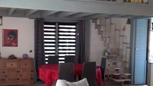 A spacious living area with a high ceiling, a staircase leading to an upper level, a dining table with chairs, and a sofa. - Maison 5/6 Pers Piscine Prive sur 6000 m2 (quissac)