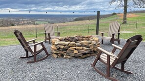 A fire pit surrounded by Adirondack chairs on a gravel surface with a scenic view in the background.