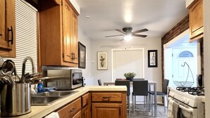 A compact kitchen with wooden cabinets, a microwave, a dishwasher, a ceiling fan, and a dining area with a table and chairs. - Redford Tranquil Retreat (Redford Charter Township)