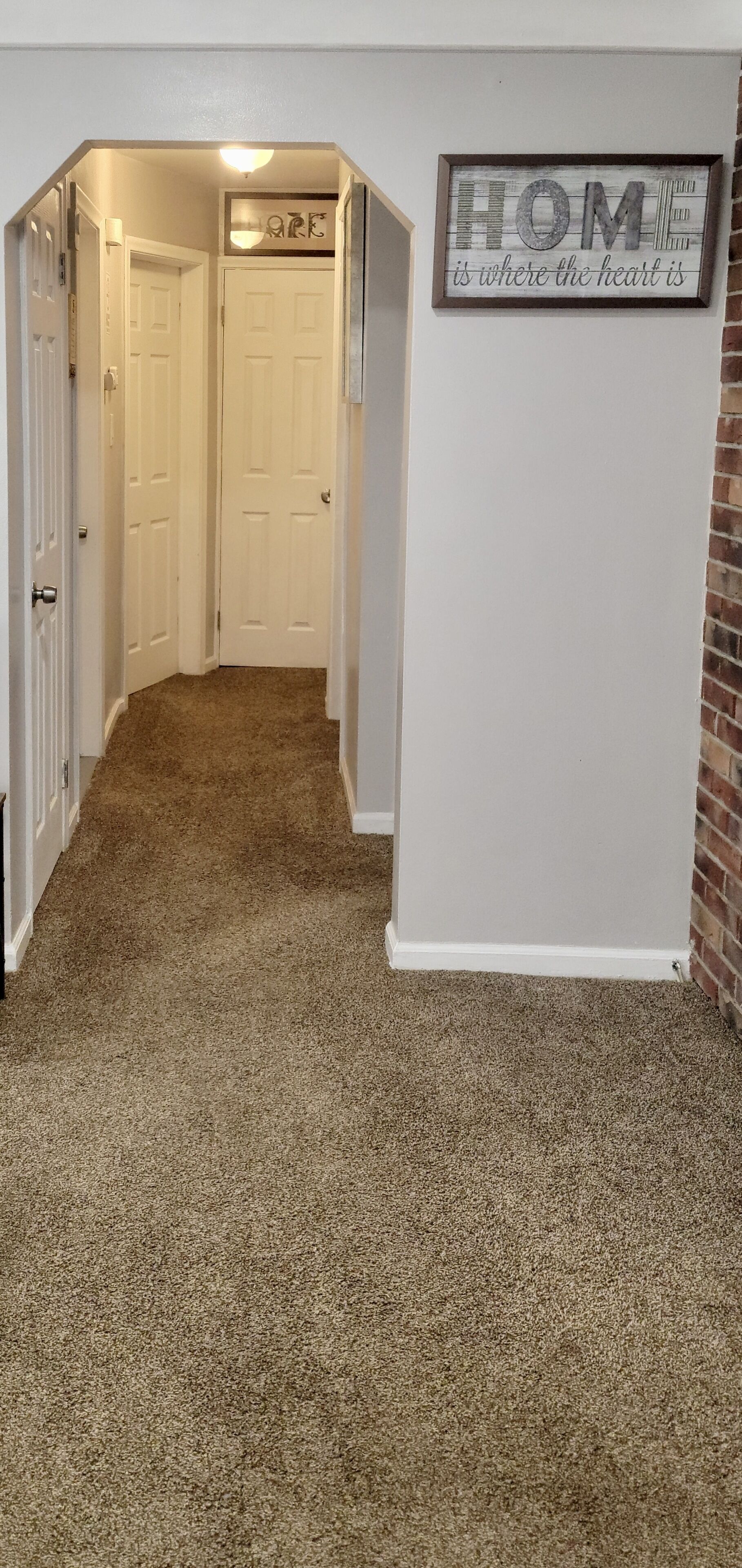 A hallway with carpeted flooring, white doors, and a 'HOME is where the heart is' sign on the wall.
