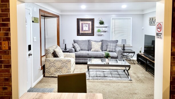 A living room with a sofa, coffee table, and a TV. There is a brick wall and a door. - Redford Tranquil Retreat (Redford Charter Township)