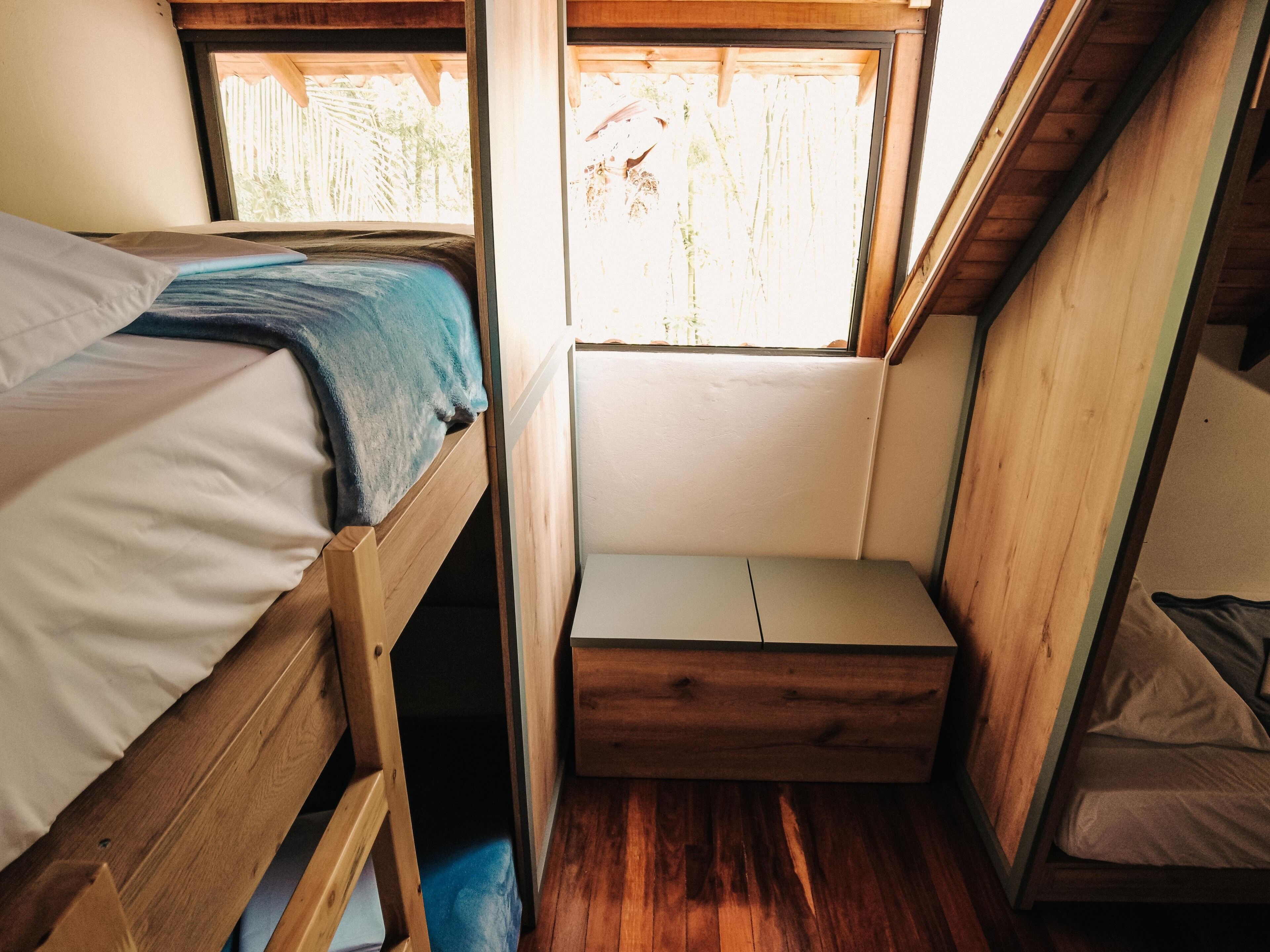 A compact room with two wooden bunk beds, a small bench, and a window providing a view of greenery outside.