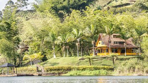 Una casa amarilla con techo de tejas, rodeada de exuberante vegetación y palmeras, cerca de una masa de agua.