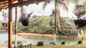 Vista desde un porche cubierto con vista a un lago, con macetas colgantes de flores y palmeras.
