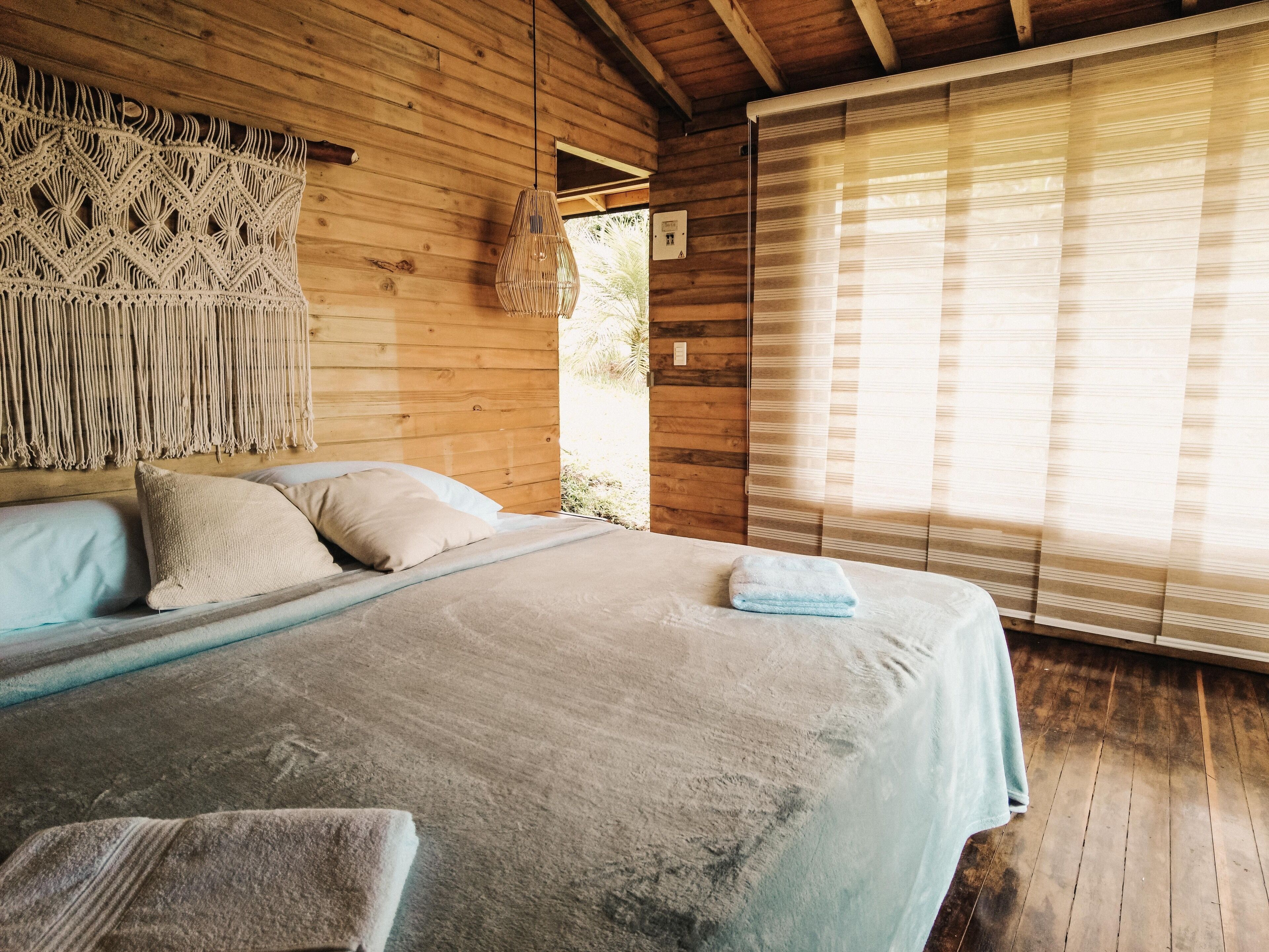 A wooden room with a bed, a macrame wall hanging, and a window with blinds.