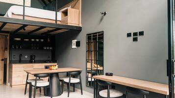 A compact, modern room with a loft bed, a kitchenette, a dining area with a wooden table and chairs, and a large mirror on the wall.