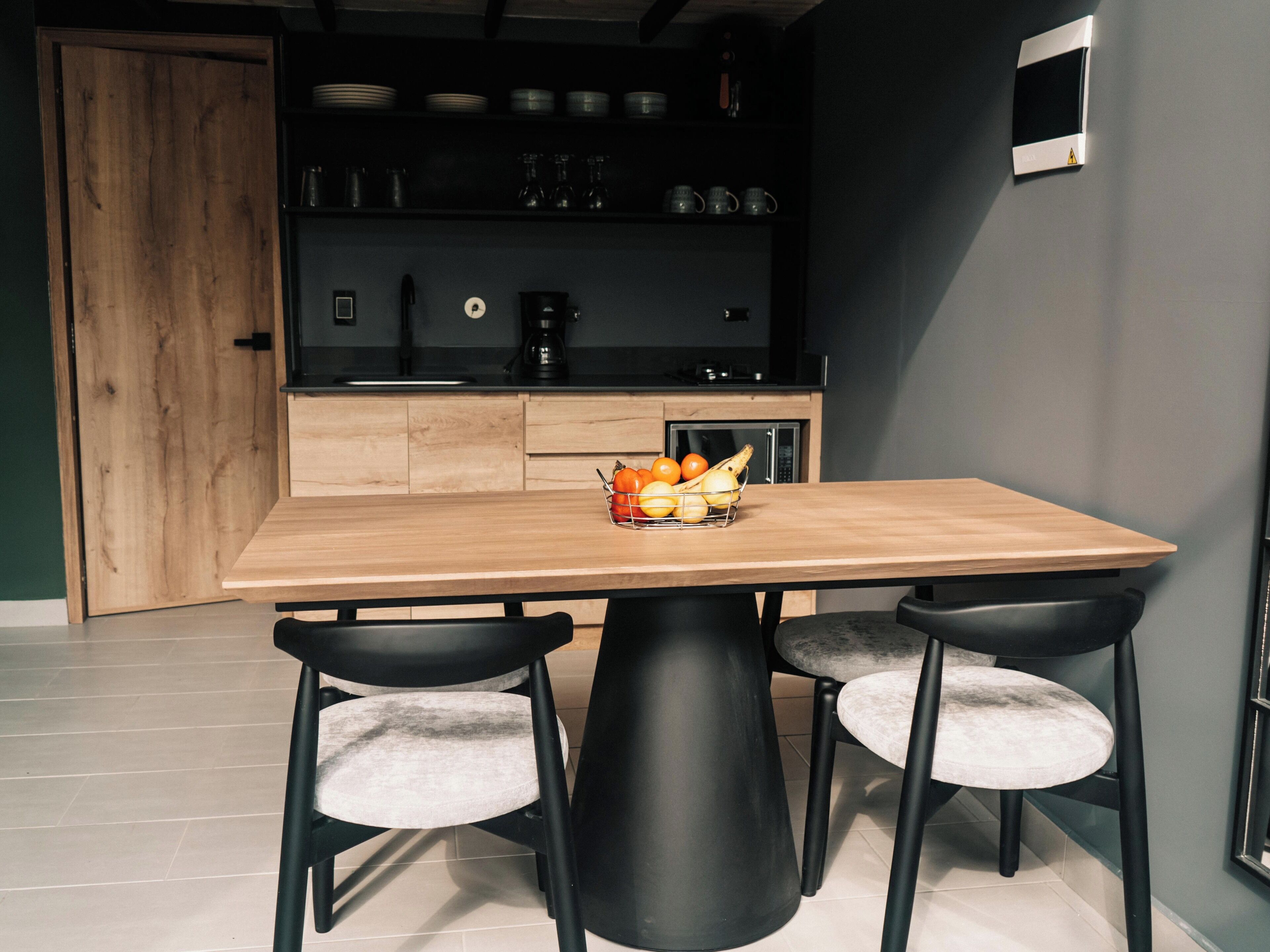 Una cocina moderna con una mesa de madera y sillas negras, un frutero sobre la mesa y un microondas al fondo.