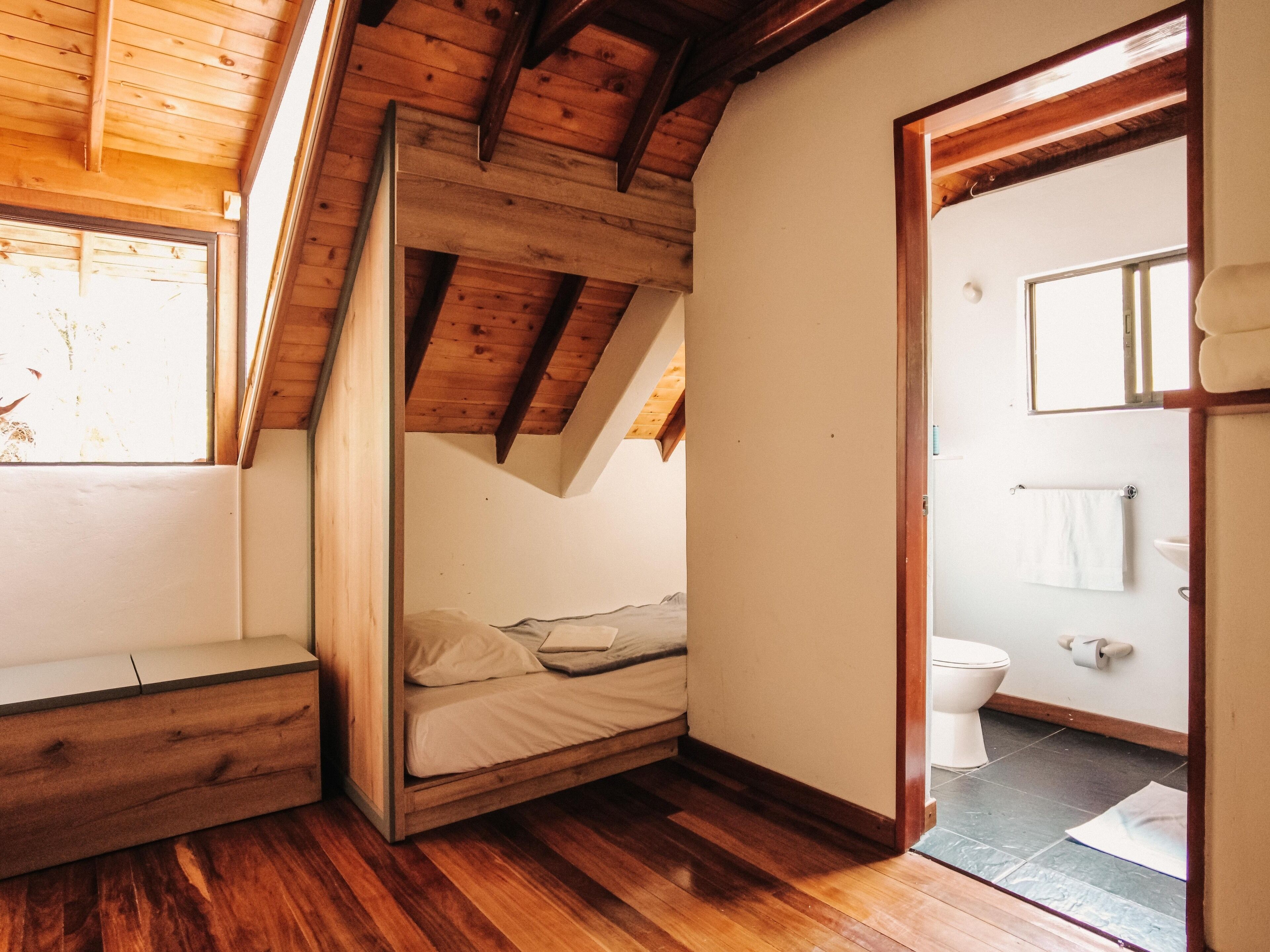 Un dormitorio pequeño y acogedor con techo de madera y una cama individual. Se ve un baño con inodoro y lavabo a través de una puerta abierta.