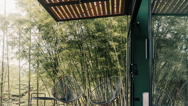 Una terraza de madera con dos sillas de alambre negro, un puerta de cristal y vista a un bosque de bambú.