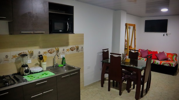 Cocina compacta con microondas, pileta y estufa. Un área de comedor con mesa de vidrio y sillas de madera. Un sofá colorido está apoyado en la pared.