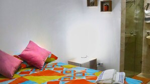 Un dormitorio con cama, almohadas, una mesita de noche, un pequeño estante con adornos, y un baño con puerta de vidrio.