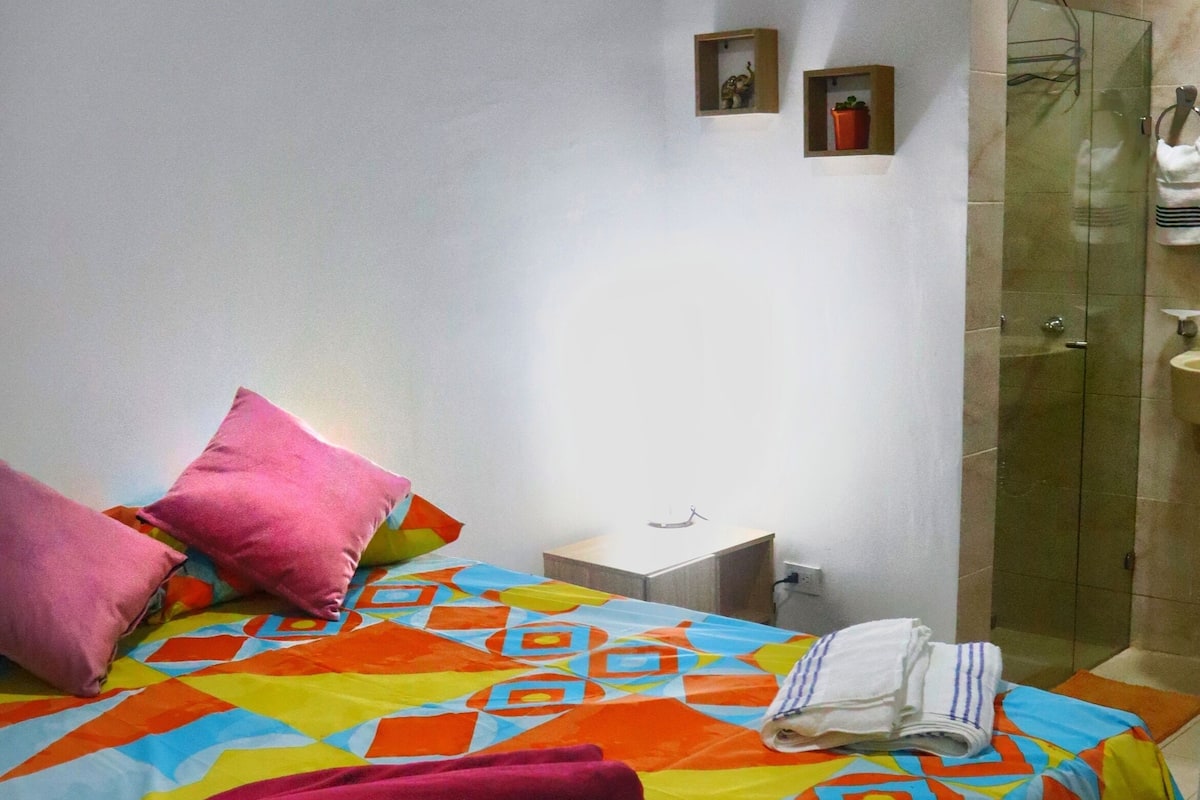 A bedroom with a bed, pillows, a bedside table, a small shelf with decorative items, and a bathroom with a glass door.