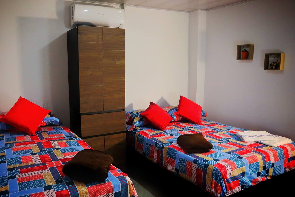 Two single beds with red pillows, a wooden wardrobe, and a wall-mounted air conditioner.