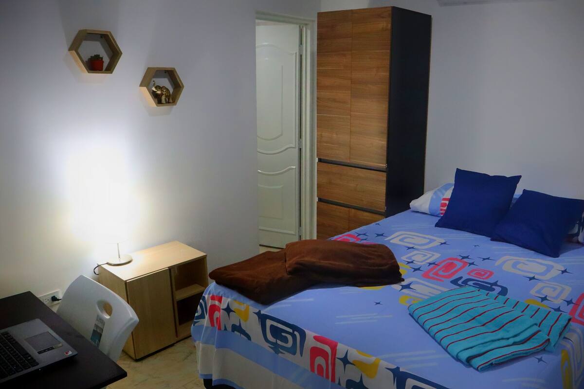 A bedroom with a bed, a desk with a laptop, a wardrobe, and two wall-mounted shelves with decorative items.