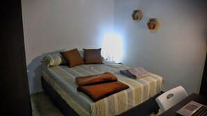 Una cama bien hecha con colcha a rayas y varios almohadones, un ordenador portátil sobre un escritorio, y dos estantes montados en la pared con objetos decorativos.