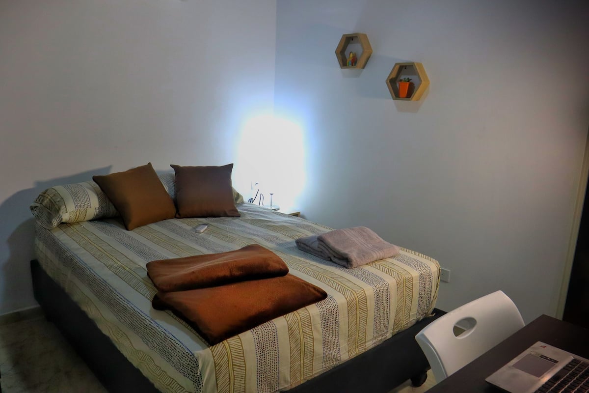 A neatly made bed with a striped bedspread and multiple pillows, a laptop on a desk, and two wall-mounted shelves with decorative items.