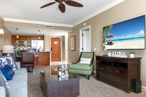 A living room with a glass coffee table, a green armchair, a sofa, and a TV displaying a beach scene.