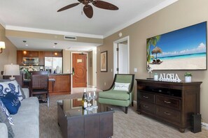 Un salon comprenant un canapé, un fauteuil vert, une table basse avec des verres à vin, un téléviseur et un tableau représentant une scène de plage.