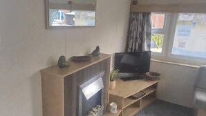 A living room with a fireplace, a flat-screen TV, and a window with curtains. - 3 Bedroom Caravan Near Mablethorpe (Louth)