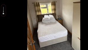 A neatly made bed with white bedding and pillows, a bedside table with a lamp, and a window with curtains. - D5 Rickardos Holiday Lets (Mablethorpe)