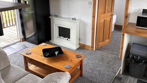 A compact living space with a sofa, a wooden coffee table, a television mounted on the wall, a fireplace, and a door leading to another room. - D5 Rickardos Holiday Lets (Mablethorpe)