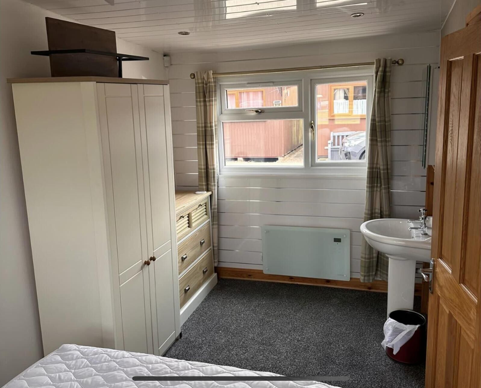 A compact bedroom with a bed, a wardrobe, a radiator, a window with curtains, and a sink.