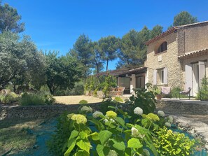 A stone building with a tiled roof, a covered patio, and a garden with a small pool and hydrangea plants. - Large Mas in the Roussillon pine forest, heated swimming pool (Gargas)