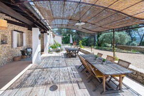 An outdoor patio with a wooden table and chairs, stone walls, and a pergola providing shade. - Large Mas in the Roussillon pine forest, heated swimming pool (Gargas)