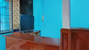 Quarto com escrivaninha de madeira, cadeira azul, televisão e janela com detalhes em ferro.