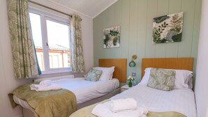 Ein Schlafzimmer mit zwei Betten, grünen Vorhängen mit Blätermotiv und zwei Wandbildern mit Blätermuster.
