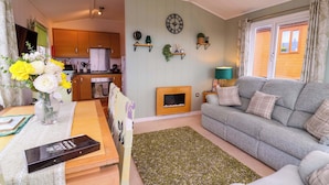 Ein gemütliches Wohnzimmer mit grauem Sofa, einem Holztisch und einem grünen Teppich. Im Hintergrund ist eine Küche mit Holzschränken und einem eingebauten Ofen zu sehen.