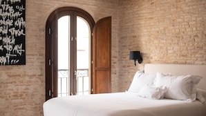 Ein Hotelzimmer mit Backsteinwand, einem großen Bett mit weißer Bettwäsche, einer Holztür und einer schwarzen Wandlampe.