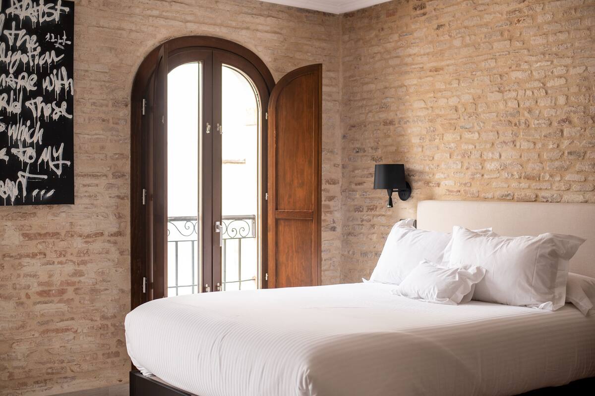 A hotel room with a brick wall, a large bed with white linens, a wooden door, and a black wall-mounted lamp.