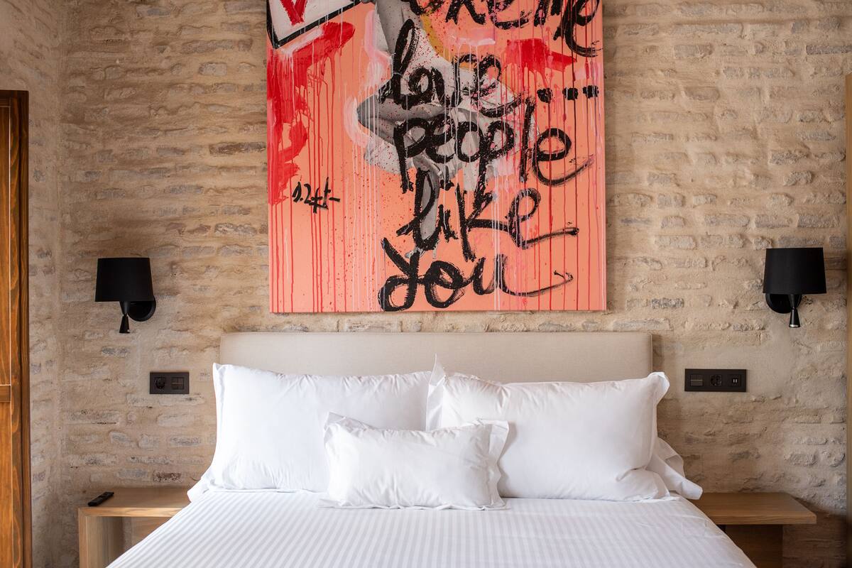 A bedroom with a brick wall, a bed with white bedding, and a piece of artwork with text.