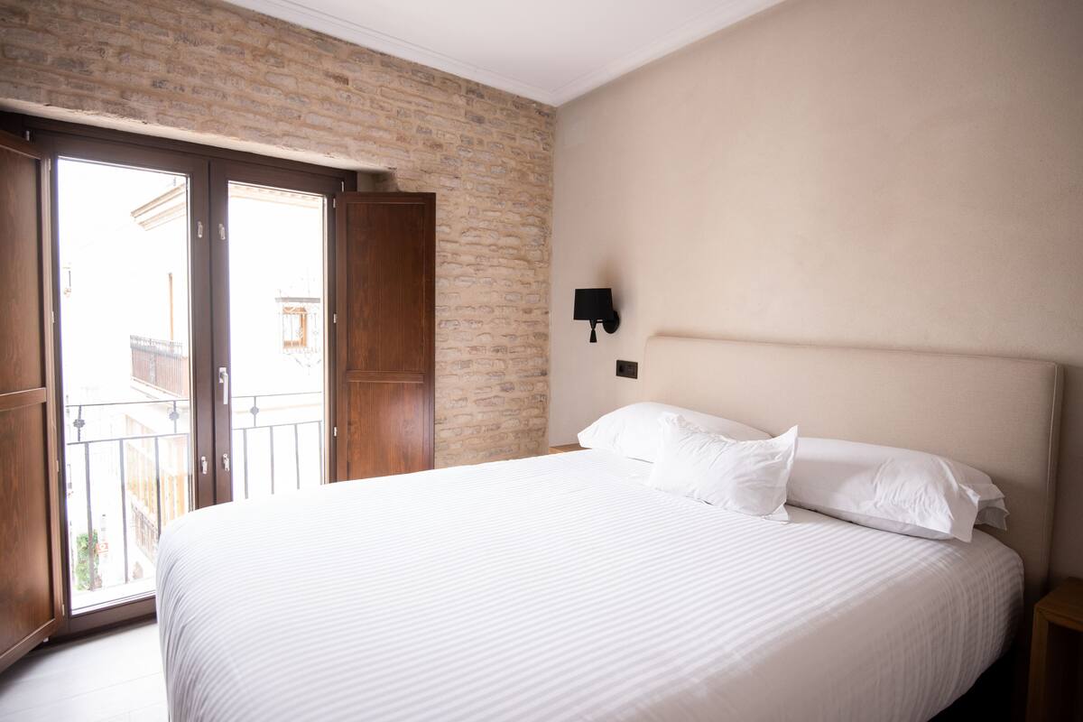 A hotel room with a large bed, a brick wall, a balcony, and a wall-mounted light fixture.