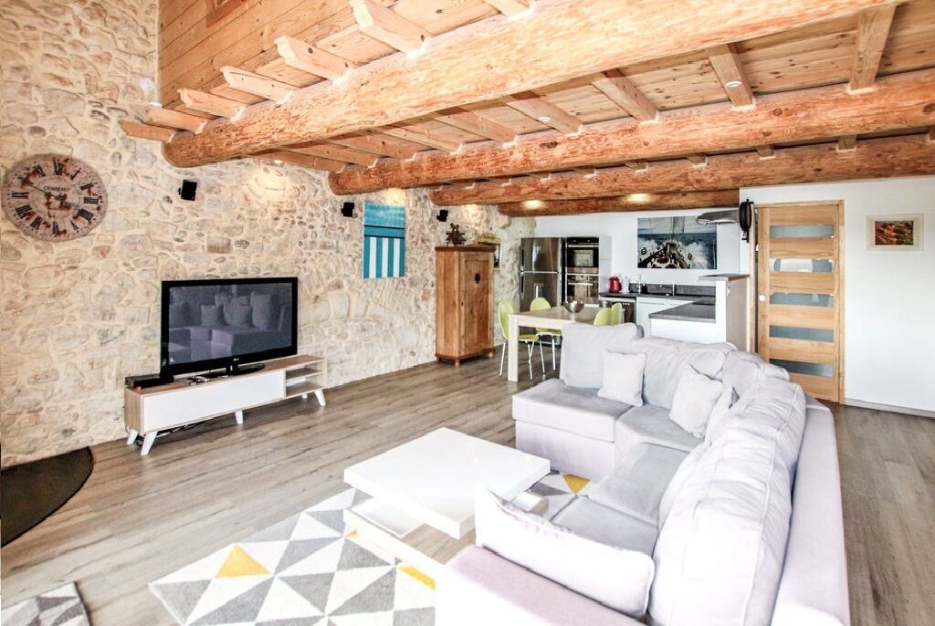 Un salon avec un mur en pierre, des poutres en bois au plafond, un téléviseur, un canapé et un coin cuisine à l’arrière-plan.