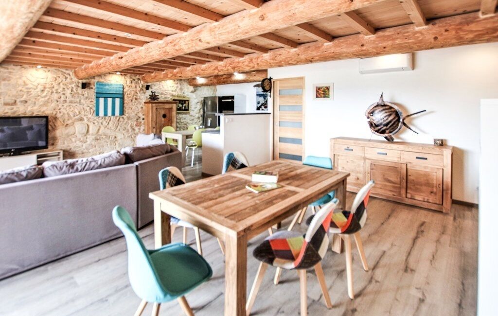 A cozy living area with a wooden dining table, colorful chairs, a stone wall, and a kitchen in the background.