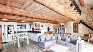 Un amplio salón con techo de madera, un sofá seccional gris grande, una mesa de comedor blanca con sillas, y una cocina al fondo.