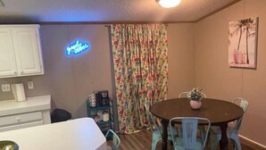 A dining area with a round table and four chairs, a neon sign, a floral curtain, and a painting on the wall.