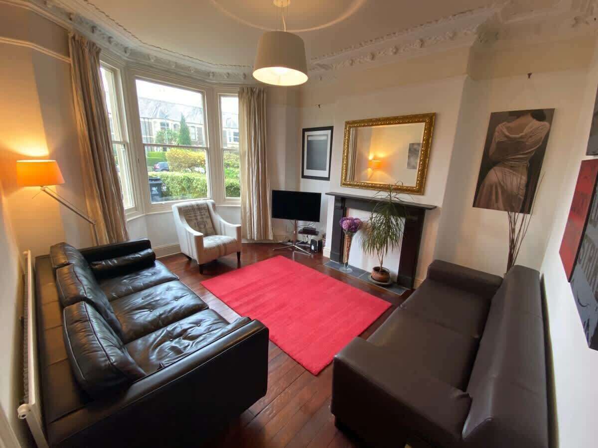 Een woonkamer met een zwarte leren bank, een rood vloerkleed, een witte fauteuil, een flatscreen-tv en een open haard.