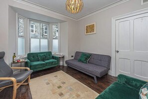 Un salon avec un canapé vert, un canapé gris, une chaise en bois, une petite table ronde et un tapis beige.