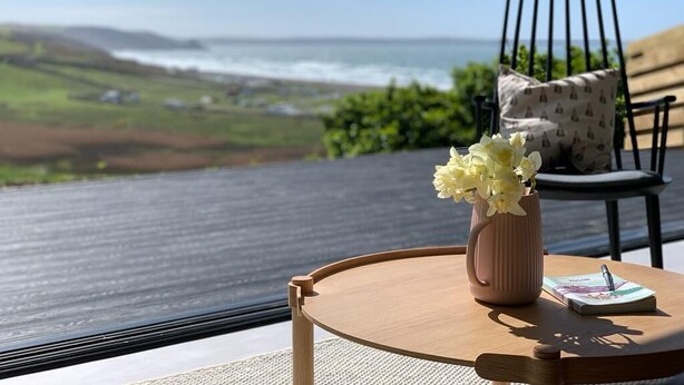A table with a vase of flowers and a notebook, overlooking a coastal landscape with a chair and cushion. - Subterranean Studio with Panoramic Views & Sauna (Pembrokeshire)