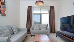Un salón con un sofá gris, una mesa de centro, un televisor de pantalla plana y un ventanal con cortinas.