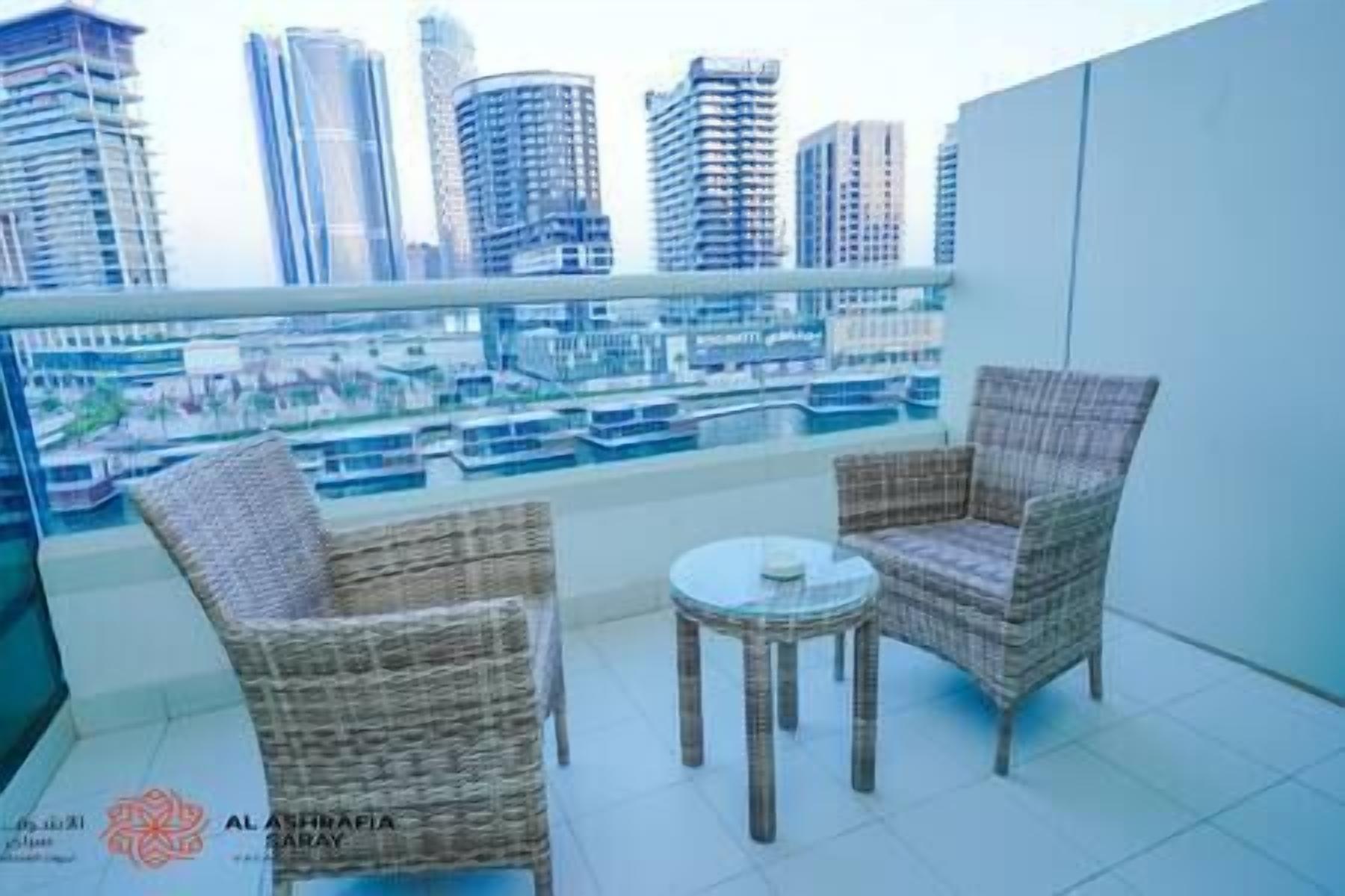 A balcony with wicker chairs and a small table, overlooking a cityscape with high-rise buildings and a river.